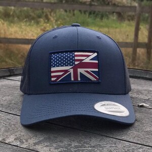 Puede incluir: Gorra de camionero azul marino con parte trasera de malla y visera curva. La gorra presenta un parche con un diseño combinado de la bandera estadounidense y británica. Una etiqueta blanca con el texto "The Authentic Snapback" está en la visera.