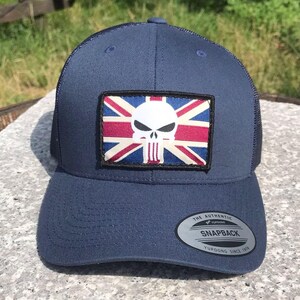 May include: A navy blue snapback cap with a mesh back. It has a patch featuring a skull design superimposed on a Union Jack flag. The cap has a curved peak and a snap closure. The tag reads "The Authentic Snapback".