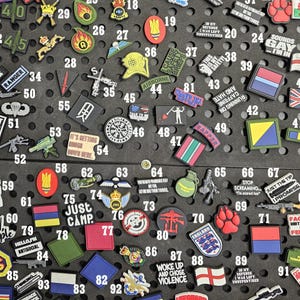 May include: A collection of various military-themed patches and pins, including flags, insignias, and humorous text. The patches feature a range of colors and designs, such as the English flag, a paw print, and the text "Sounds Gay I'm In".