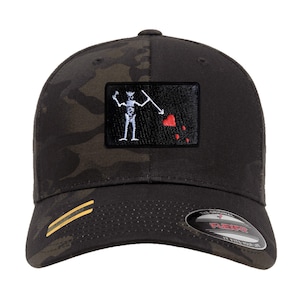 Op de afbeelding: Zwarte en camouflage baseballcap met een Flexfit-ontwerp. De pet heeft een rechthoekige patch met een wit skelet met een speer en een rood hart. De klep heeft een gouden streep.
