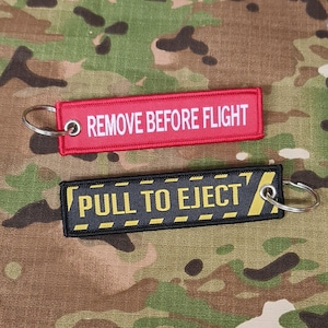May include: Two embroidered keychains. One is red with white text that reads "REMOVE BEFORE FLIGHT". The other is black with yellow text that reads "PULL TO EJECT". Both have metal key rings.