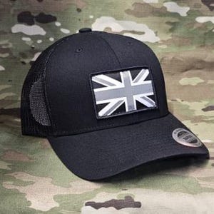 May include: Black trucker hat with a mesh back and a curved brim. The hat features a rectangular patch with a gray and white Union Jack flag design. A small, silver sticker is on the brim. The hat is set against a camouflage background.