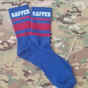 May include: A pair of blue socks with red and white stripes. The word "SAPPER" is printed in white block letters across the top of each sock. The socks are displayed on a camouflage background.