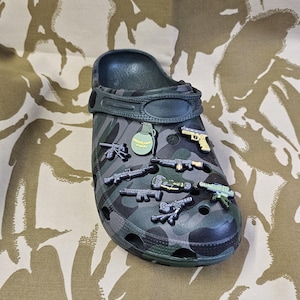 Puede incluir: Un zueco de camuflaje verde oscuro adornado con dijes temáticos de armas. Los dijes incluyen granadas, pistolas y rifles en miniatura. El diseño del zapato incorpora un patrón de camuflaje en tonos verdes y grises.