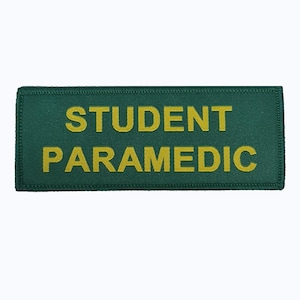Puede incluir: Parche rectangular verde con las palabras "STUDENT PARAMEDIC" en letras amarillas. El parche tiene un borde cosido, probablemente para un uniforme o equipo médico.