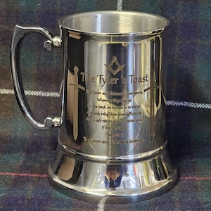 Pode incluir: Um caneco de prata polida com uma alça grande e uma base alargada. A caneca apresenta texto e símbolos gravados, incluindo as palavras "The Tyler's Toast" e um emblema maçónico. Duas espadas estão gravadas na lateral. O fundo é um padrão xadrez.