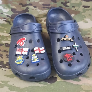 May include: Two navy blue clogs decorated with charms, including flags and military emblems. One clog features the text "Royal Navy Commando", the other "Navy Veteran". The clogs have multiple holes for ventilation.