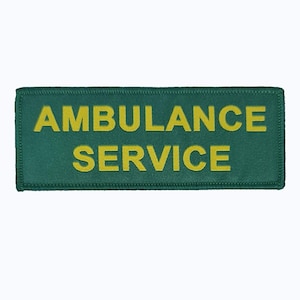 Puede incluir: Un parche rectangular verde con las palabras "AMBULANCE SERVICE" en letras amarillas. El parche tiene un borde cosido, probablemente para la identificación del uniforme.