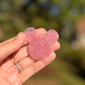 May include: A pink, crystal-covered Mickey Mouse-shaped accessory. The accessory is held in a hand, with a blurred green and brown background. The accessory is made of many small, sparkling crystals.