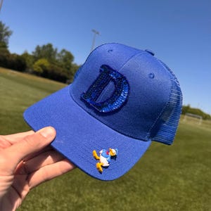 May include: A royal blue trucker hat with a mesh back and a curved brim. The hat features a large, sequined "D" on the front and a Donald Duck charm. The hat is held up against a green field and blue sky.