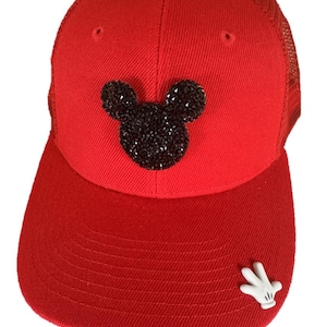 May include: Red trucker hat with a black rhinestone Mickey Mouse head design on the front. The hat has a curved brim and a white hand emblem on the side. The back of the hat is mesh.