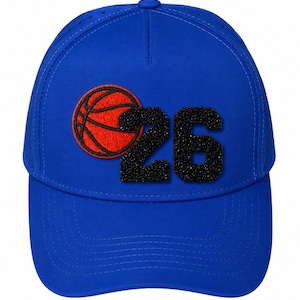 Peut inclure: Une casquette de baseball bleu roi ornée d'un graphique de ballon de basket orange texturé à côté du chiffre 26 noir. La casquette présente une calotte structurée avec des panneaux latéraux perforés et une visière incurvée au design épuré.