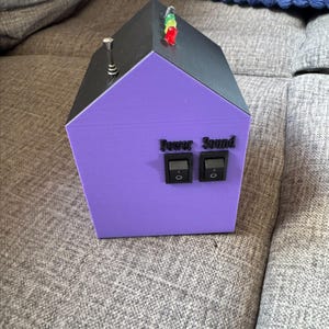 May include: A house-shaped electronic device with a purple front and black roof. It features two black rocker switches labeled Power and Sound, three small LED lights in green, yellow, and red on the roof, and a small metallic antenna on the side.