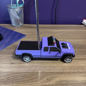 May include: A purple toy pickup truck with a black bed, silver wheels, and a tall antenna. The truck has a black roof and the text "POWER" on the front. The toy is sitting on a wooden surface.