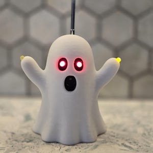 May include: A white ghost-shaped figurine with a smooth matte finish. The ghost features two glowing red circular eyes and a black oval-shaped mouth. Small yellow lights are positioned at the tips of each raised arm, and a thin metal rod extends upward.