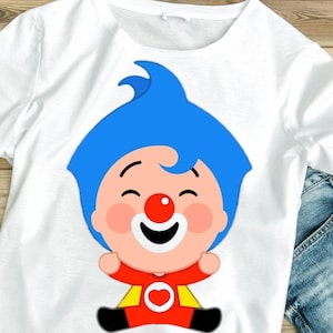 May include: White t-shirt featuring a cartoon character with blue hair, a red nose, and a smiling face. The character is wearing a red and yellow outfit with a heart emblem. The shirt is displayed on a wooden surface with a pair of blue jeans.