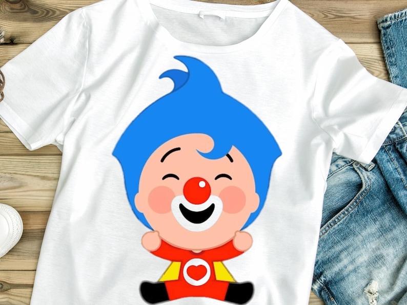 May include: White t-shirt featuring a cartoon character with blue hair, a red nose, and a smiling face. The character is wearing a red and yellow outfit with a heart emblem. The shirt is displayed on a wooden surface with a pair of blue jeans.