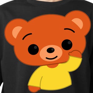 May include: Black t-shirt featuring a cartoon bear with orange fur, black eyes, and a yellow shirt. The bear is waving with its right paw. The design is centered on the shirt.