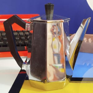 Può includere: Una caffettiera argentata con manico e coperchio neri. La caffettiera ha un design geometrico ed è posta su uno sfondo colorato giallo, blu e rosso.