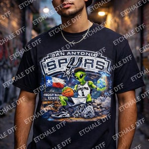 May include: Black t-shirt featuring a graphic of an alien wearing a Spurs basketball jersey, holding a basketball and a planet. The shirt also displays the San Antonio Spurs logo and the Texas flag.