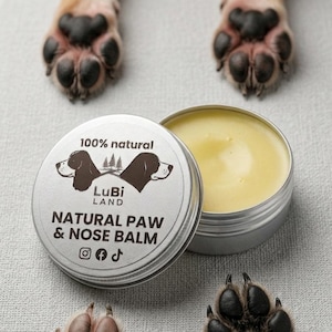 May include: A silver tin of natural paw and nose balm, open to reveal a yellow balm. The lid is labelled "100% natural" and "LuBi Land NATURAL PAW & NOSE BALM". Dog paws are visible in the image.