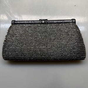May include: A rectangular clutch bag covered in small, dark grey metallic beads. The bag features a rigid, dark silver-toned metal frame along the top edge with an intricate, embossed cross-hatch pattern and a central clasp mechanism.