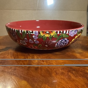 May include: A large, round ceramic bowl with a deep red interior and exterior. The exterior features a vibrant, hand-painted floral pattern with yellow, purple, and red flowers, green leaves, and a decorative white and red striped border at the rim.
