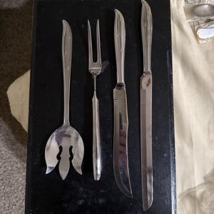 May include: A set of four silver-toned stainless steel serving utensils featuring a decorative star pattern on the handles. The set includes a wide salad fork, a two-pronged meat fork, a curved carving knife, and a long, straight slicing knife.