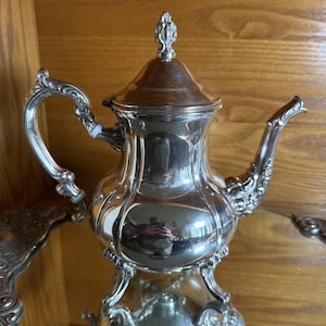 Puede incluir: Una tetera de tono plateado pulido con un asa curva, un pico largo y una tapa extraíble con un remate superior. El cuerpo de la tetera tiene una forma redondeada y multifacética con detalles decorativos ornamentados en la base y el asa.