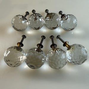 May include: Eight clear crystal ball cabinet knobs with faceted surfaces. Each knob has a metal stem and a decorative brass-colored base. The knobs are arranged in two rows of four on a white surface.