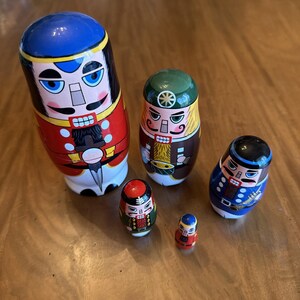 May include: A set of five nesting dolls painted as nutcracker figures in various uniforms. The largest doll wears a red coat and blue hat, while the others feature green, blue, and red outfits with painted details like drums and trumpets.