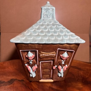 May include: A ceramic cookie jar shaped like a schoolhouse with a light blue speckled roof. The brown exterior features two elf-like figures flanking a central door, with a sign above the door clearly displaying the text SCHOOLHOUSE in raised letters.