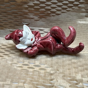 Könnte beinhalten: Eine Keramikfigur eines liegenden Elfen mit einem glänzend dunkelroten Körper und einem weißen Gesicht. Der Elf hat spitze Ohren, geschlossene Augen und leuchtend rote Lippen. Er liegt auf der Seite mit einem angewinkelten Bein und glatter Oberfläche.