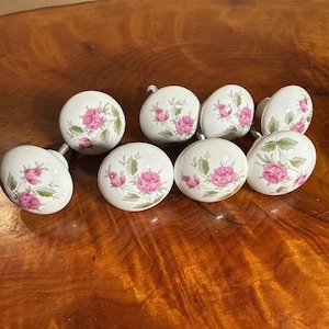 May include: A set of seven round white ceramic cabinet knobs, each featuring a painted floral design of pink roses with green leaves and stems. The knobs have a smooth, glossy finish and include metal hardware attachments for installation.