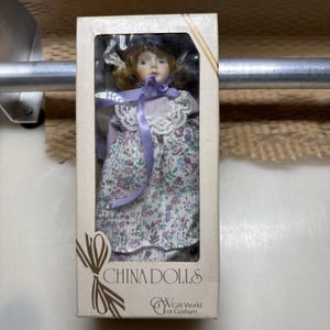 May include: A porcelain doll with brown hair, wearing a lavender dress with a white lace collar and a purple ribbon tie, encased in a rectangular cardboard box labeled CHINA DOLLS and Gift World of Gorham with a decorative bow graphic.