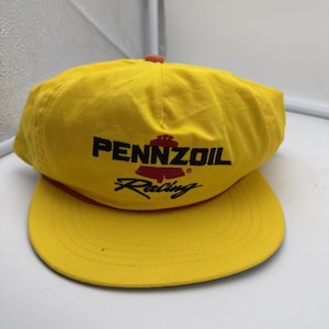 May include: A bright yellow baseball cap featuring the black text PENNZOIL above a red bell-shaped logo and the word Racing in black script. The cap has a red cord trim along the base of the crown and a matching yellow brim with stitching.