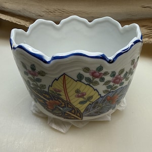 May include: A white ceramic bowl with a scalloped rim and a blue painted edge. The exterior features a colourful pattern of stylised fan shapes in yellow, blue, and dark red, accented with small pink flowers and green leaves on thin stems.