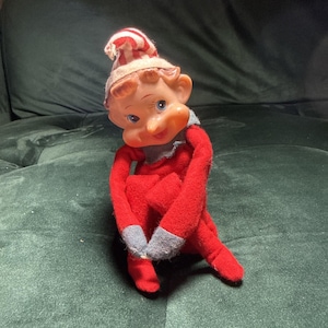 May include: A small elf doll with a plastic face, blue eyes, and a pointed nose. The doll wears a red and white striped hat, a red felt body, and grey felt mittens and boots. The elf is posed in a seated position with its arms wrapped around its knees.