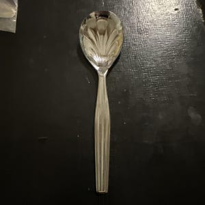 Puede incluir: Una cuchara de metal plateado con un cuenco que presenta un patrón decorativo de concha en relieve y un mango caracterizado por líneas verticales acanaladas. La superficie pulida refleja la luz, resaltando los detalles texturizados.