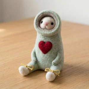 May include: A needle-felted creature with white fur and black eyes, wearing a light sage green hooded suit featuring a dark red heart on the chest. The figure is in a seated position with small yellow string bows tied around each of its white feet.