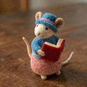May include: A needle-felted mouse figurine wearing a blue hat with a pink band, a blue jumper, and pink trousers. The mouse is holding an open red book in its paws. The figure has light beige fur, small dark eyes, and a long, thin, light-coloured tail.