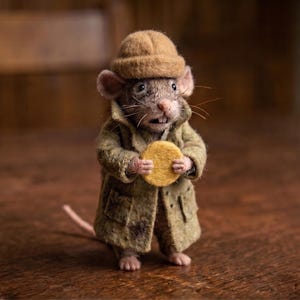Puede incluir: Una figura de ratón de fieltro con un sombrero de fieltro color canela y una gabardina verde oliva texturizada. El ratón tiene orejas rosadas, una cola larga y sostiene un pequeño disco amarillo redondo entre sus patas delanteras, de pie.