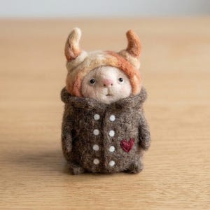 May include: A needle-felted creature figurine wearing a tan and orange horned hood and a dark brown coat. The coat features two vertical rows of small white dots and a single small red heart embroidered on the lower right side of the garment.