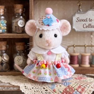 May include: A handcrafted white mouse figurine wearing a dress with a floral pattern, a hat, and a collar. The mouse has pink ears and a pink nose. The background includes jars filled with buttons and beads, spools of thread, and a sign that reads "Sweet to Cherish Collection."
