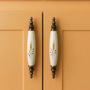 May include: Two cabinet pulls with a wheat stalk design. The pulls have a white, oval-shaped center with a gold wheat stalk and bronze-colored hardware. The pulls are mounted on a light orange cabinet.