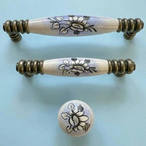 May include: A set of three cabinet hardware pieces: two handles and one knob. The handles and knob feature a cream base with a floral design in black and white, with light blue accents. The hardware has a bronze finish.
