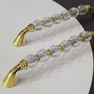 May include: Two cabinet pulls featuring a series of clear, faceted, spherical glass beads separated by polished gold-tone metal rings. Each handle terminates in a flared, fan-shaped gold-tone metal base with a smooth, reflective finish.