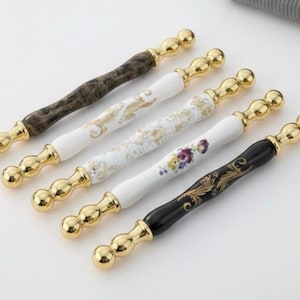May include: A collection of five decorative cabinet handles. Each handle features a gold-colored, bulbous end and a central section with varying designs. The central sections include a marbled brown, white with gold filigree, white with floral accents, and black with gold filigree.