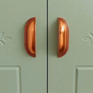 May include: Close-up of two copper-colored cabinet handles with a half-moon design. The handles are mounted on a light green cabinet door. The cabinet door has a recessed panel and a floral design carved into the upper right corner.
