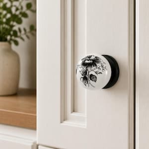 May include: A round white ceramic cabinet knob featuring a detailed black floral illustration of a blooming flower with leaves. The knob is mounted on a black circular base and is attached to a white paneled cabinet door with vertical grooves.